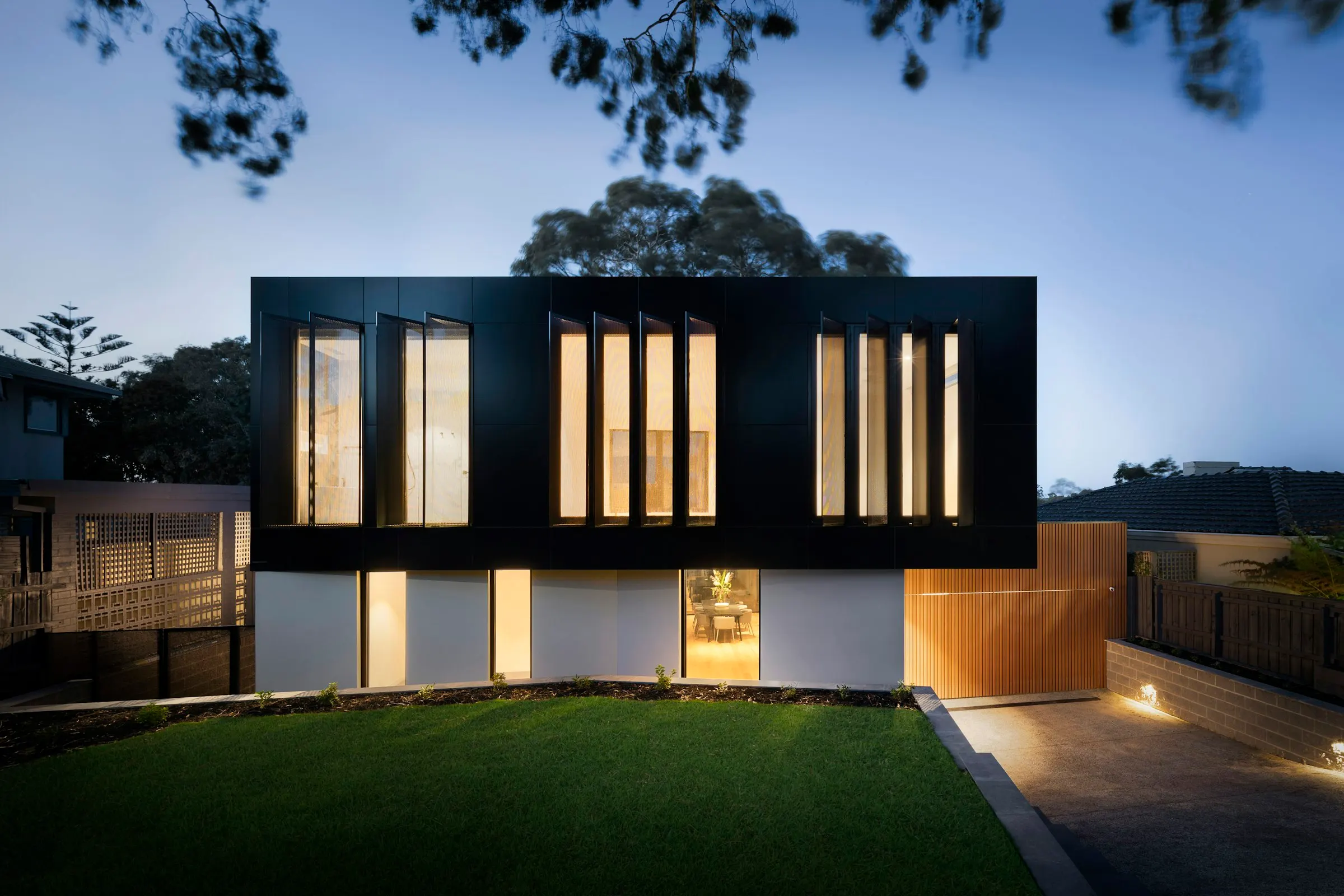 Dark timber house at dusk with warmly lit vertical windows above a glazed ground floor